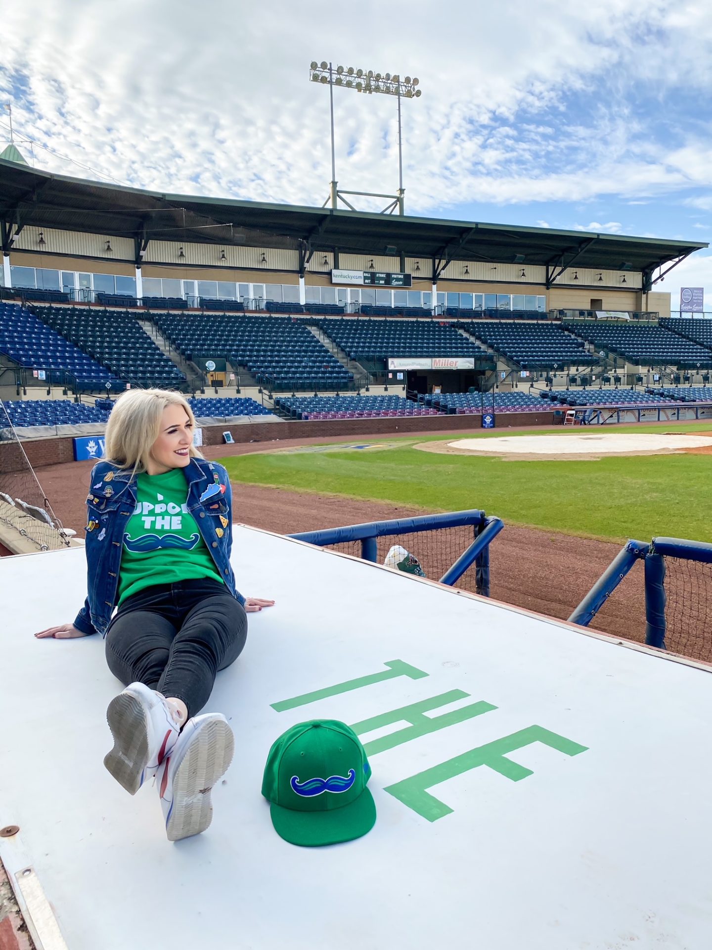 blonde girl sitting on top of the dugout at a ballpark